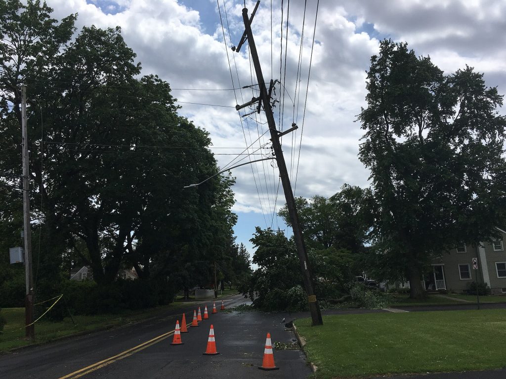 A utility pole leaning over a residential street with the overhead power lines pulled down out of position after a storm, traffic cones on the road and mature trees on both sides.